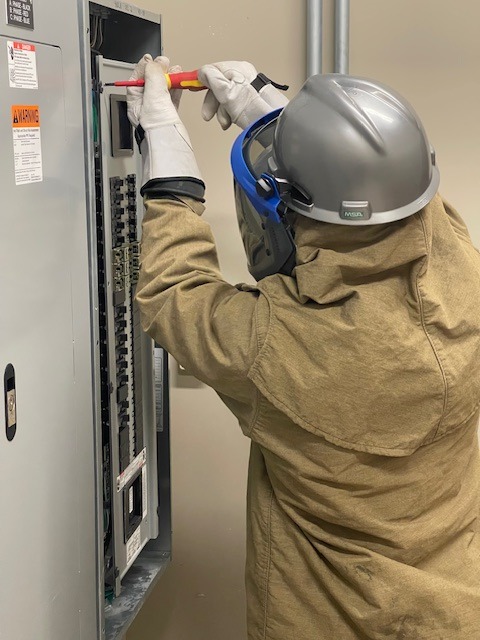 A technician in an arc-rated suit and face shield working inside an energized panel