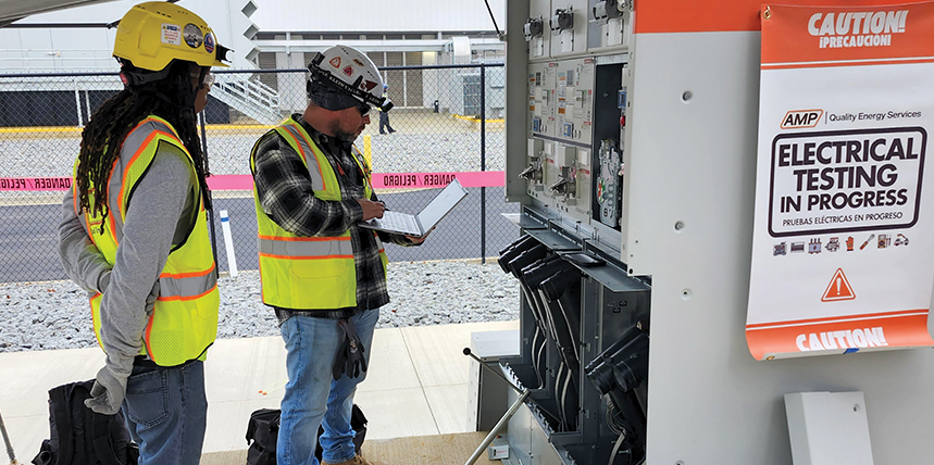 Two AMP technicians testing energized switchgear behind an electrical testing in progress barrier